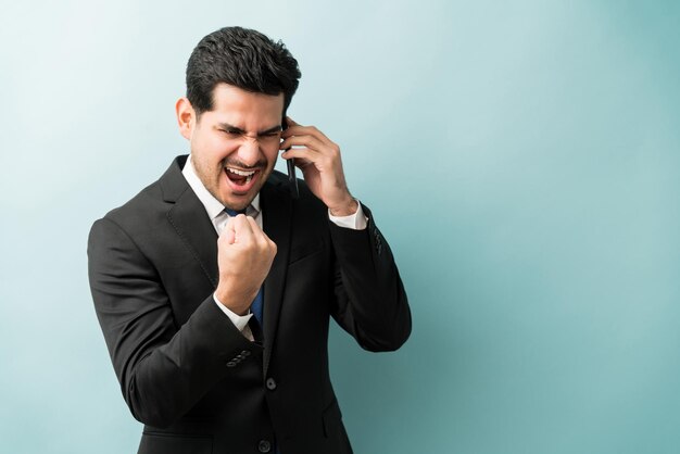 Emocionado hombre de negocios guapo hablando por teléfono móvil apretando el puño después de escuchar buenas noticias contra el fondo azul