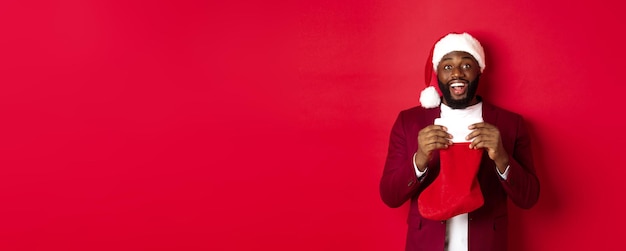 Emocionado hombre negro abre calcetín de navidad con regalos y dulces sonriendo feliz de pie en santa hat a
