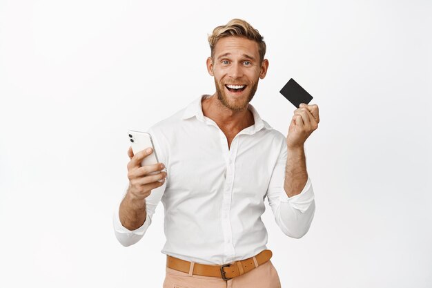 Emocionado hombre sonriente usando la aplicación de teléfono inteligente y sacudiendo la tarjeta de crédito en la mano ganando dinero en el teléfono móvil de pie sobre fondo blanco.