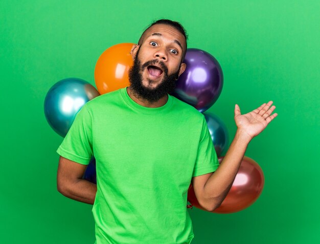 Emocionado joven afroamericano con camiseta verde de pie delante de globos extendiendo la mano aislada en la pared verde