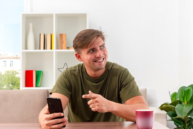 Emocionado joven apuesto rubio se sienta a la mesa con la taza sosteniendo y apuntando al teléfono mirando al lado