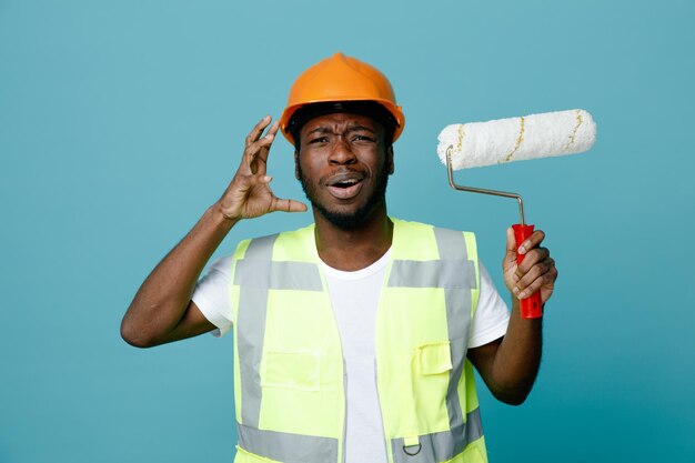 Emocionado joven constructor afroamericano en uniforme con cepillo de rodillos aislado sobre fondo azul.