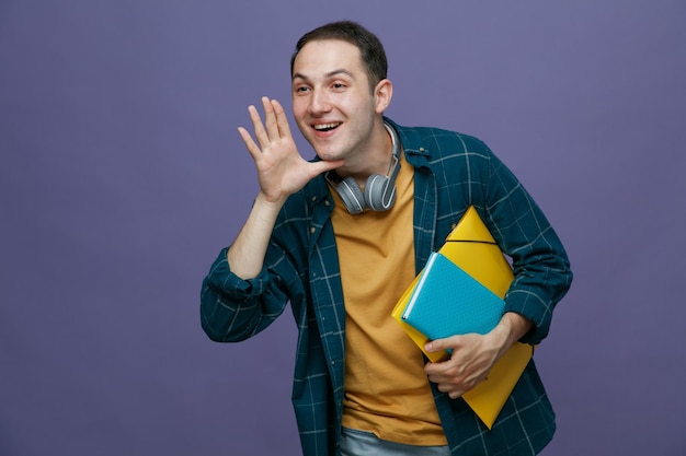 Emocionado joven estudiante masculino usando audífonos alrededor del cuello sosteniendo un cuaderno de carpetas manteniendo la mano cerca de la boca mirando al costado llamando a alguien aislado en un fondo morado