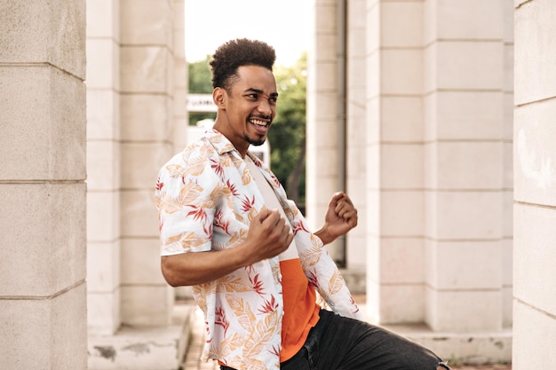 Emocionado joven feliz en camiseta colorida y camisa floral se regocija y se mueve de buen humor al aire libre