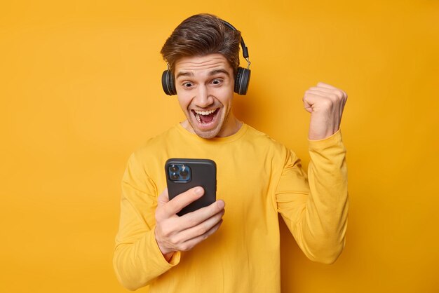 Emocionado joven jugador juega videojuegos aprieta el puño con triunfo mira la pantalla del teléfono inteligente celebra ganar un nuevo nivel usa auriculares en los oídos aislados sobre fondo amarillo Sí, lo hice