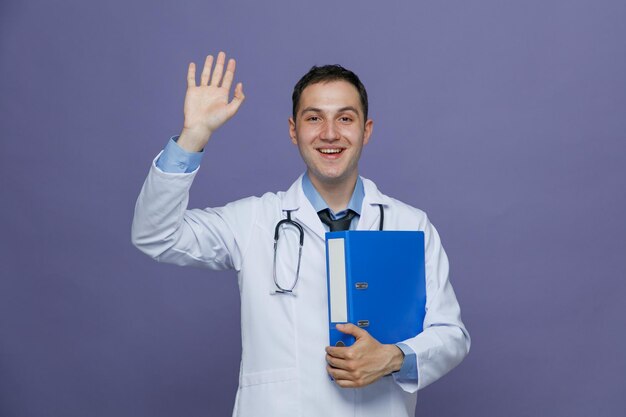 Emocionado joven médico masculino con bata médica y estetoscopio alrededor del cuello sosteniendo una carpeta abrazándola mirando a la cámara mostrando un gesto de hola aislado en un fondo morado