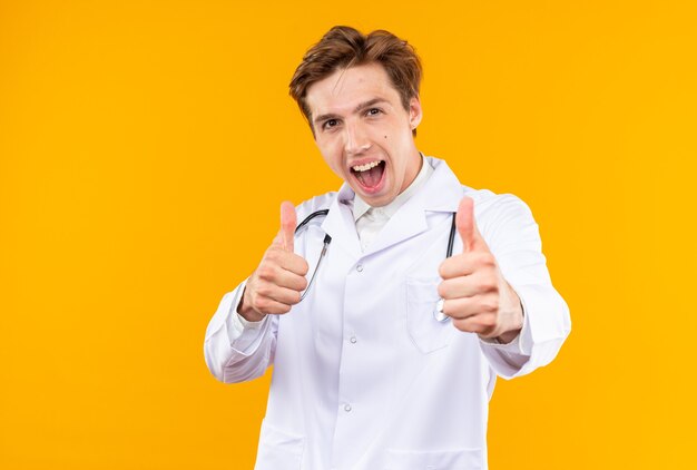 Emocionado joven médico vistiendo bata médica con estetoscopio mostrando el pulgar hacia arriba aislado en la pared naranja