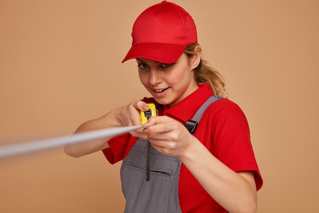 Emocionado joven trabajador de la construcción con gorra y uniforme estirando el medidor de cinta hacia la cámara