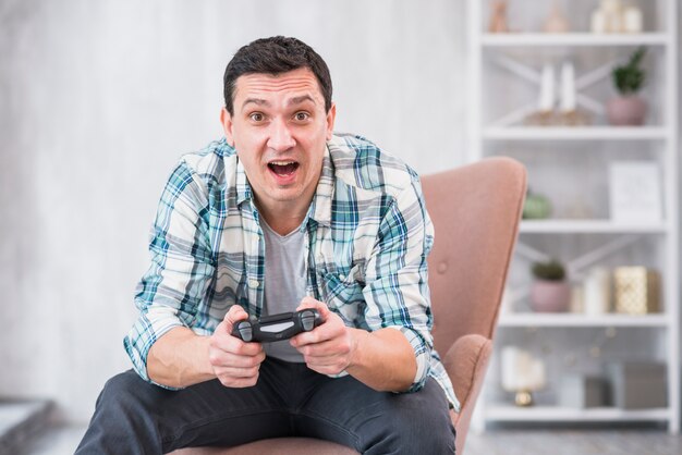 Emocionado joven varón sentado en el sillón y jugando con gamepad