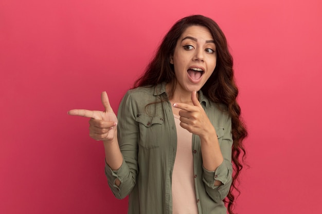 Emocionado mirando al lado hermosa joven vestida con puntos de camiseta verde oliva en el lado aislado en la pared rosa con espacio de copia