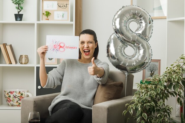 Emocionado mostrando el pulgar hacia arriba hermosa niña en el día de la mujer feliz con tarjeta de felicitación sentada en un sillón en la sala de estar