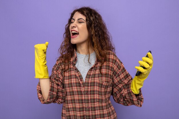 Emocionado mostrando sí gesto joven mujer de la limpieza con guantes sosteniendo el teléfono aislado en la pared púrpura