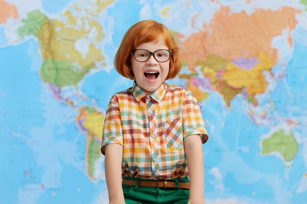 Emocionado niño pequeño pelirrojo con gafas grandes y camisa a cuadros, abriendo la boca con alegría mientras estaba de pie en la sala de clase, feliz de ver a sus padres y volver a casa. Niño inteligente