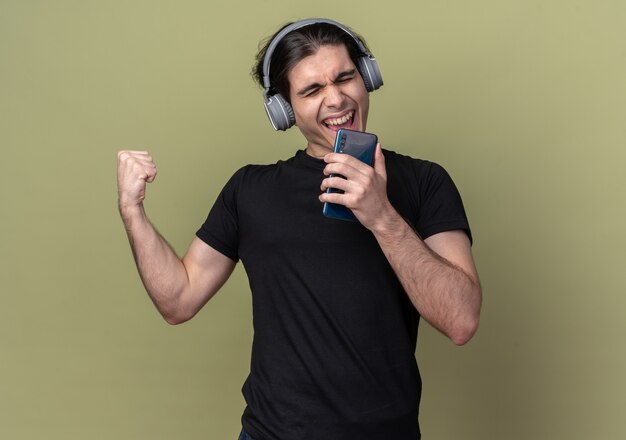 Emocionado con los ojos cerrados joven guapo con camiseta negra y auriculares sosteniendo el teléfono y canta aislado en la pared verde oliva