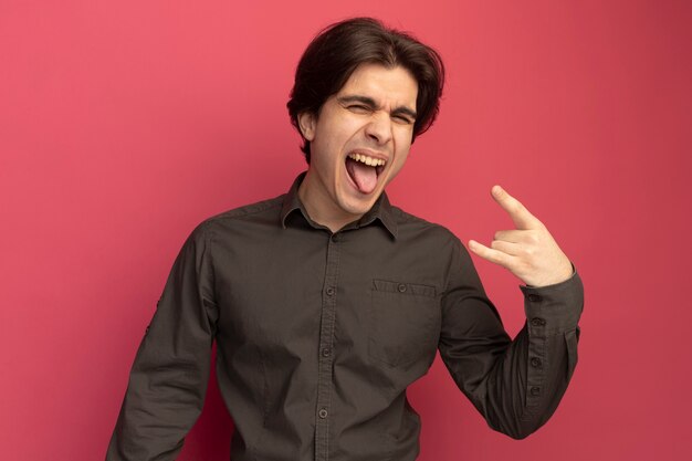 Emocionado con los ojos cerrados joven guapo con camiseta negra mostrando la lengua y haciendo gesto de cabra aislado en la pared rosa