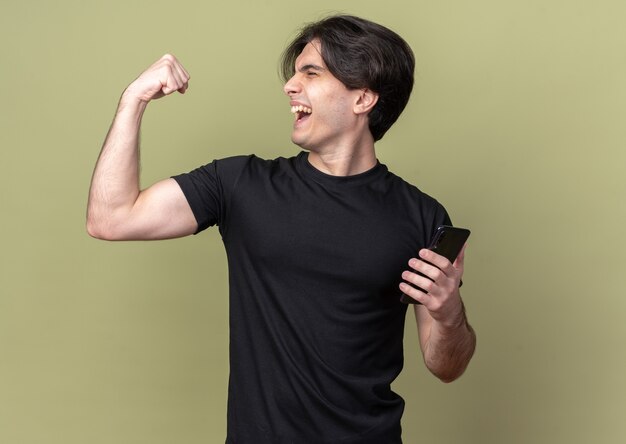 Emocionado con los ojos cerrados joven guapo con camiseta negra sosteniendo el teléfono haciendo un gesto fuerte aislado en la pared verde oliva