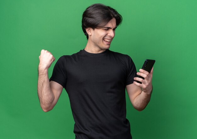 Emocionado con los ojos cerrados joven guapo con camiseta negra sosteniendo el teléfono mostrando sí gesto aislado en la pared verde