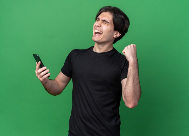 Emocionado con los ojos cerrados joven guapo con camiseta negra sosteniendo el teléfono mostrando sí gesto aislado en la pared verde