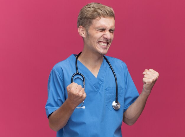 Emocionado con los ojos cerrados joven médico con uniforme de médico con estetoscopio mostrando gesto sí aislado en la pared rosa