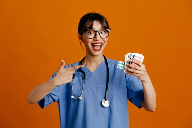 Emocionado sosteniendo píldoras joven doctora vistiendo uniforme fith estetoscopio aislado sobre fondo naranja