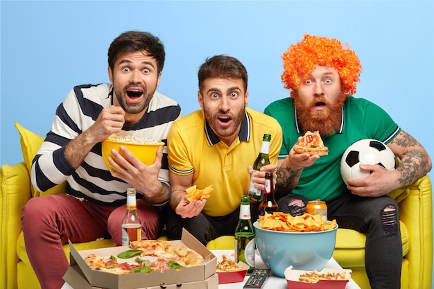 Emocionados tres amigos varones concentrados en la pantalla del televisor, miran el partido de fútbol con gran interés, posan en el sofá de la espaciosa sala de estar, comen palomitas de maíz