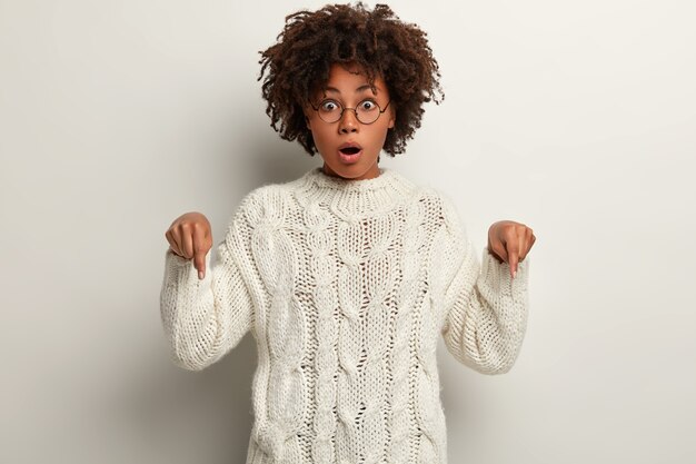 Emocional atractiva mujer afroamericana apunta hacia abajo con expresión de asombro impresionado, usa lentes ópticos y un jersey, modelos sobre una pared blanca. Concepto de publicidad. Mira hacia abajo.