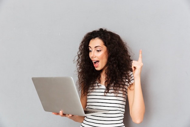 Foto gratuita emocional dama con cabello rizado con cuaderno plateado para encontrar información útil en internet gesticulando con el dedo índice sobre la pared gris