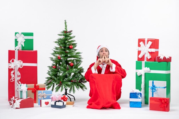 Emocional emocionado joven vestido como Papá Noel con regalos y árbol de Navidad decorado haciendo gesto de silencio sobre fondo blanco.