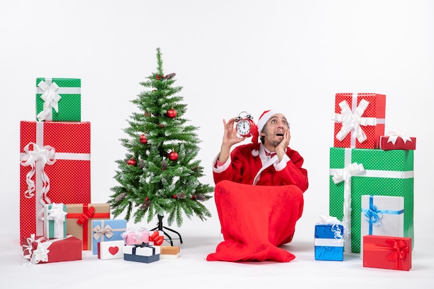 Emocional feliz papá noel sorprendido mirando arriba sentado en el suelo y mostrando el reloj cerca de regalos y árbol de Navidad decorado sobre fondo blanco.