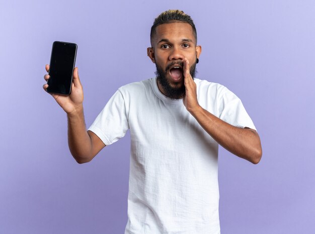 Emocional joven afroamericano en camiseta blanca mostrando smartphone gritando con la mano sobre la boca de pie sobre fondo azul.