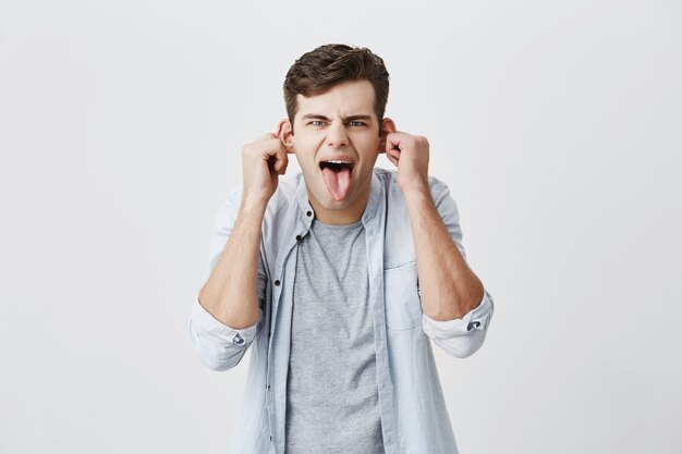 Emocional joven estudiante masculino en camisa azul haciendo muecas, burlándose, haciendo muecas, sacando orejas, sacando la lengua, mirando con expresión loca y estúpida en su rostro.