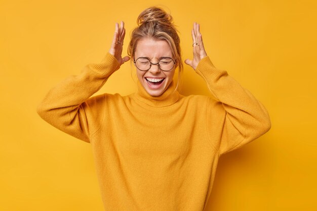 Emocional joven europea exclama en voz alta mantiene las manos levantadas sobre la cabeza mantiene los ojos cerrados reacciona ante algo asombroso lleva gafas redondas puente casual aislado sobre fondo amarillo