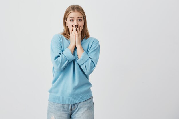 Emocional joven mujer en suéter azul y jeans que parece asustada tocando su barbilla con ambas manos mirando con gran sorpresa en la cámara sorprendida después de escuchar noticias horrorizadas sobre sus amigos