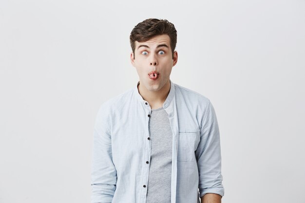 Emocional joven varón caucásico con camisa azul haciendo muecas, mirando a los ojos, haciendo caras tontas, sacando la lengua, mirando con expresión loca y estúpida en su rostro