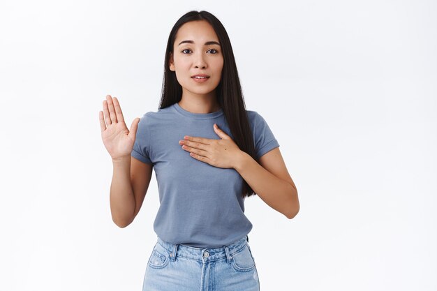 Emocional, linda chica asiática promete decir solo la verdad, poner una mano en el corazón y levantar el brazo mientras jura, hacer una declaración honesta, hacer un juramento o promesa, decir un voto mientras mira a la cámara, pared blanca