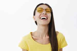 Foto gratuita emocional mujer atractiva despreocupada y optimista con cabello oscuro en gafas de sol amarillas redondas levantando la cabeza mientras se ríe