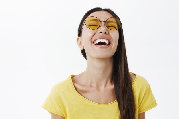 Emocional mujer atractiva despreocupada y optimista con cabello oscuro en gafas de sol amarillas redondas levantando la cabeza mientras se ríe