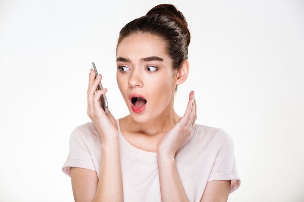 Emocional mujer con cabello castaño en moño expresando sorpresa mientras tiene conversación móvil