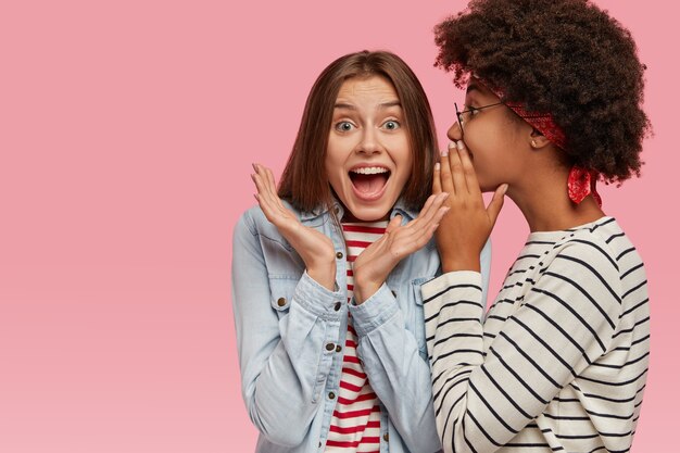 Emocional mujer europea se da la mano y exclama en voz alta mientras escucha los rumores de su mejor amiga. Mujer afroamericana susurra un secreto al oído de sus compañeros