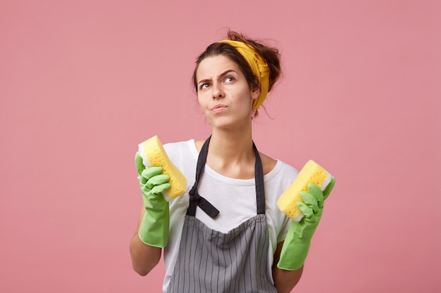 Foto gratuita emocional mujer joven con delantal y guantes de goma obsesionada con la limpieza, sosteniendo esponjas en ambos brazos mientras limpia en la cocina. concepto de higiene, tareas domésticas y limpieza.