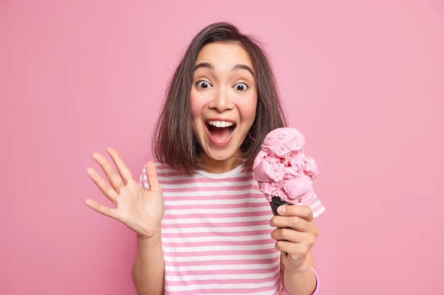 Emocional mujer morena exclama en voz alta mantiene la palma levantada come delicioso helado reacciona ante algo inesperado ha sorprendido mirada alegre viste camiseta a rayas casual aislada sobre pared rosa