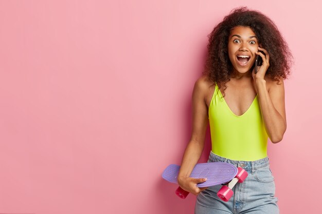 Emocional mujer de piel oscura habla por teléfono celular, le cuenta noticias a un amigo, se viste con un traje de verano a la moda, sostiene el longboard, se para contra la pared rosa, copia el espacio a un lado. Concepto de estilo de vida