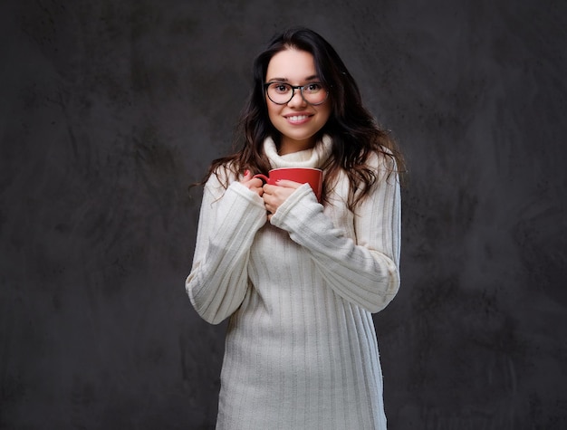 Emocional y sonriente mujer morena vestida con un cálido suéter de cuello alto sostiene una gran taza de café roja sobre fondo gris.