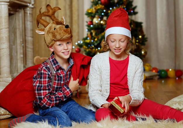 Emocionante chico lindo con sombrero de ciervo de Navidad y chica feliz sostiene una caja de regalo en una habitación decorada con Navidad.