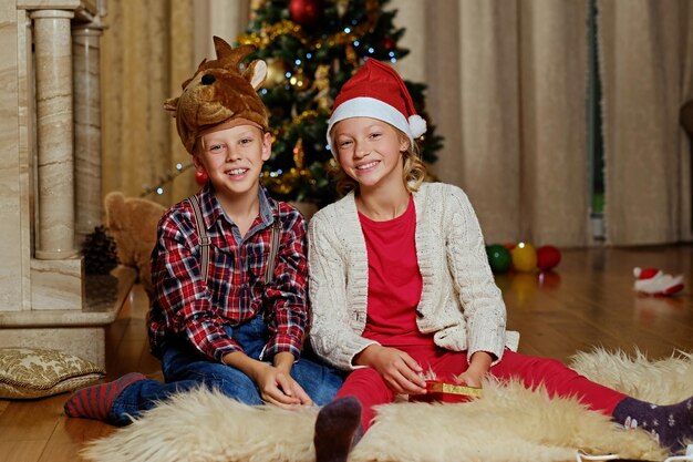 Emocionante chico lindo con sombrero de ciervo de Navidad y chica feliz sostiene una caja de regalo en una habitación decorada con Navidad.
