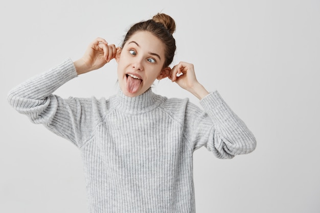 Emociones divertidas de hilarante hembra tocando sus orejas y sacando la lengua. Mujer caucásica con cabello castaño en moño tirando de la cara haciendo muecas. Alegría, concepto de diversión