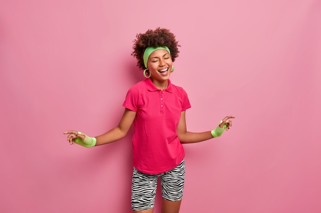 Emociones felices y concepto de estilo de vida. Alegre mujer deportiva de piel oscura baila con alegría, vestida con ropa deportiva, siendo enérgica