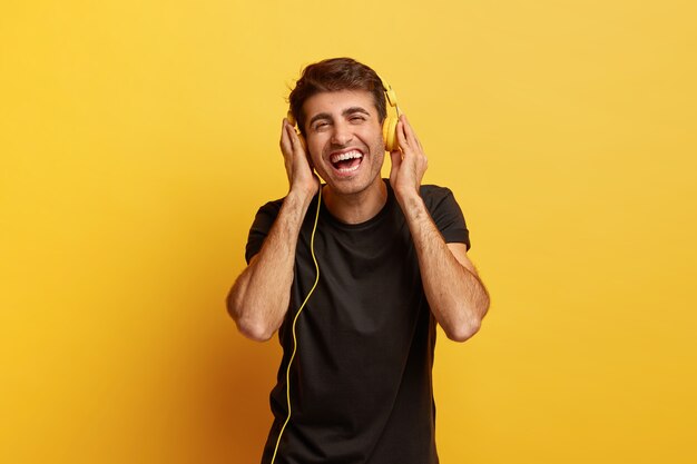 Emociones humanas positivas y concepto de diversión. Feliz alegre hombre caucásico mantiene ambas manos en auriculares modernos, sonríe ampliamente