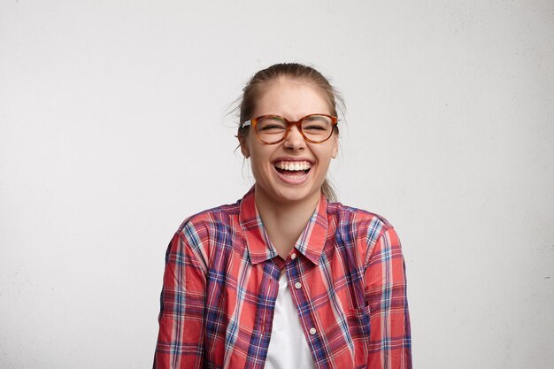 Emociones humanas positivas. Retrato de estudio de una mujer joven relajada y despreocupada con una gran sonrisa que usa anteojos cerrando los ojos con fuerza mientras se ríe a carcajadas de los buenos chistes, divirtiéndose con amigos en el interior