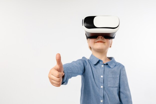 Emociones más frescas. Niña o niño en jeans y camisa con gafas de casco de realidad virtual aisladas sobre fondo blanco de estudio. Concepto de tecnología de punta, videojuegos, innovación.